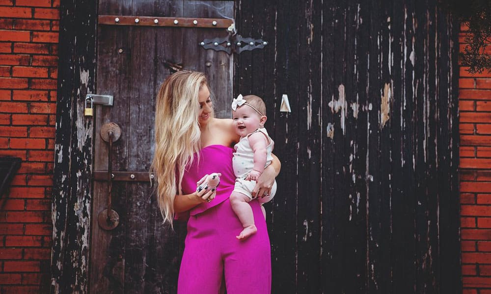 woman in pink jumper, holding baby, in front of a wall